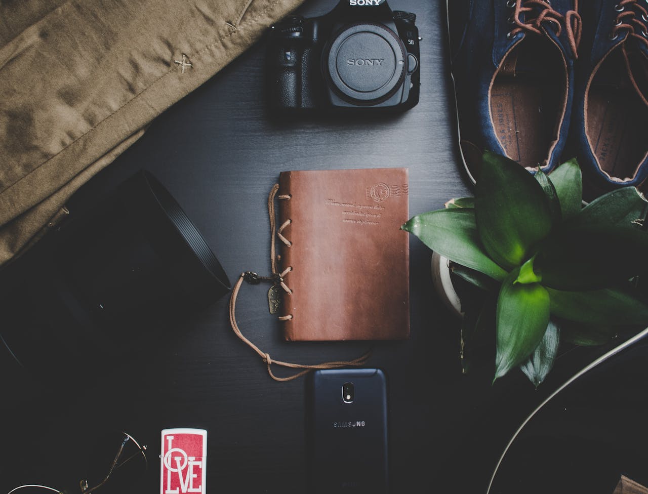 pexels-photo-1447263 Flatlay featuring travel essentials including a camera, notebook, smartphone, and plant on a dark surface.
